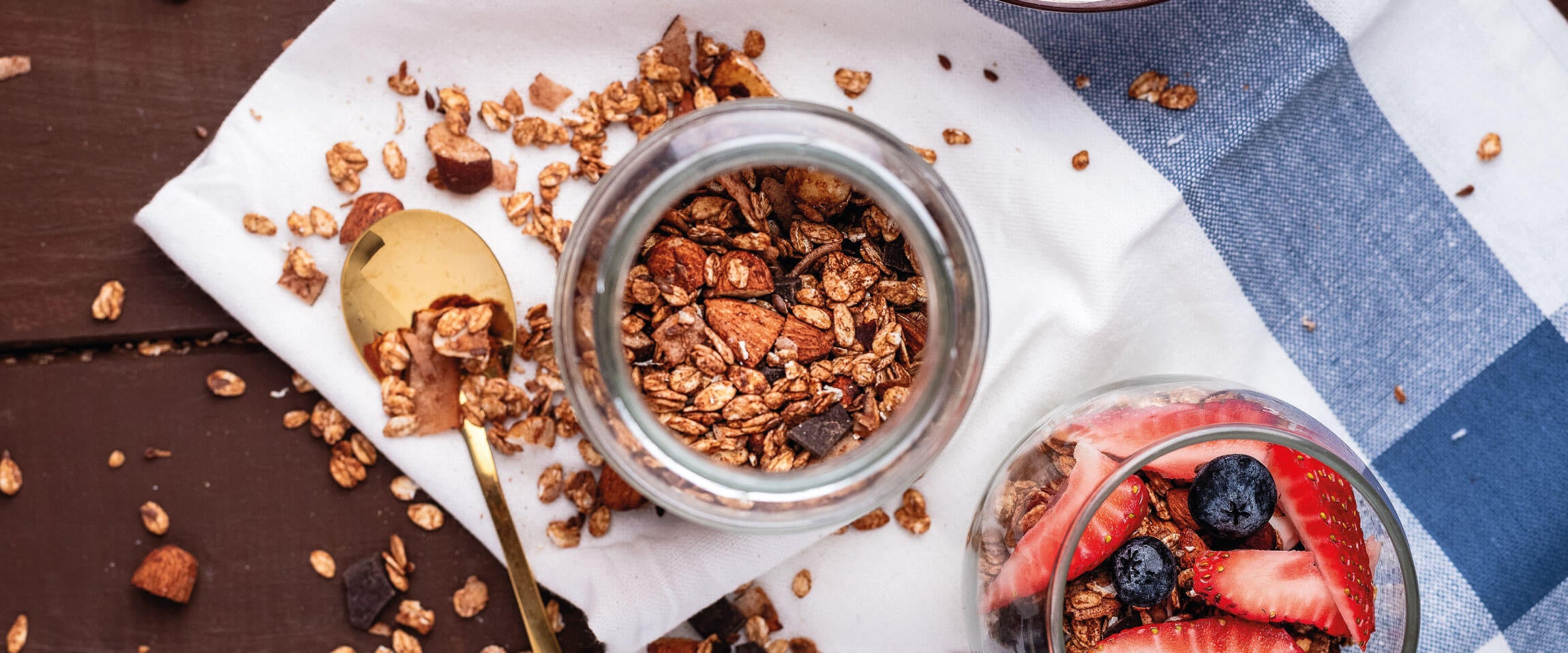 Se ve un envase con granola de chocolate y fresas frescas, sobre una manta blanca y una cuchara dorada.