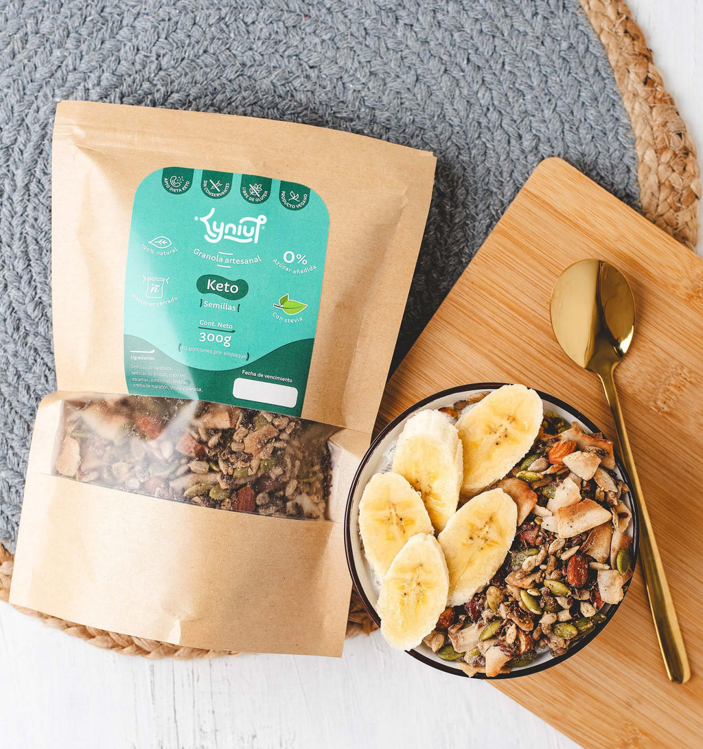 Bolsa de granola artesanal keto semillas Kyniut de 300g con empaque kraft y etiqueta verde, junto a un bowl servido con rodajas de banano sobre una tabla de madera y una cuchara dorada en una toma cenital.