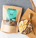 Bolsa de granola artesanal keto semillas Kyniut de 300g con empaque kraft y etiqueta verde, junto a un bowl servido con rodajas de banano sobre una tabla de madera y una cuchara dorada en una toma cenital.
