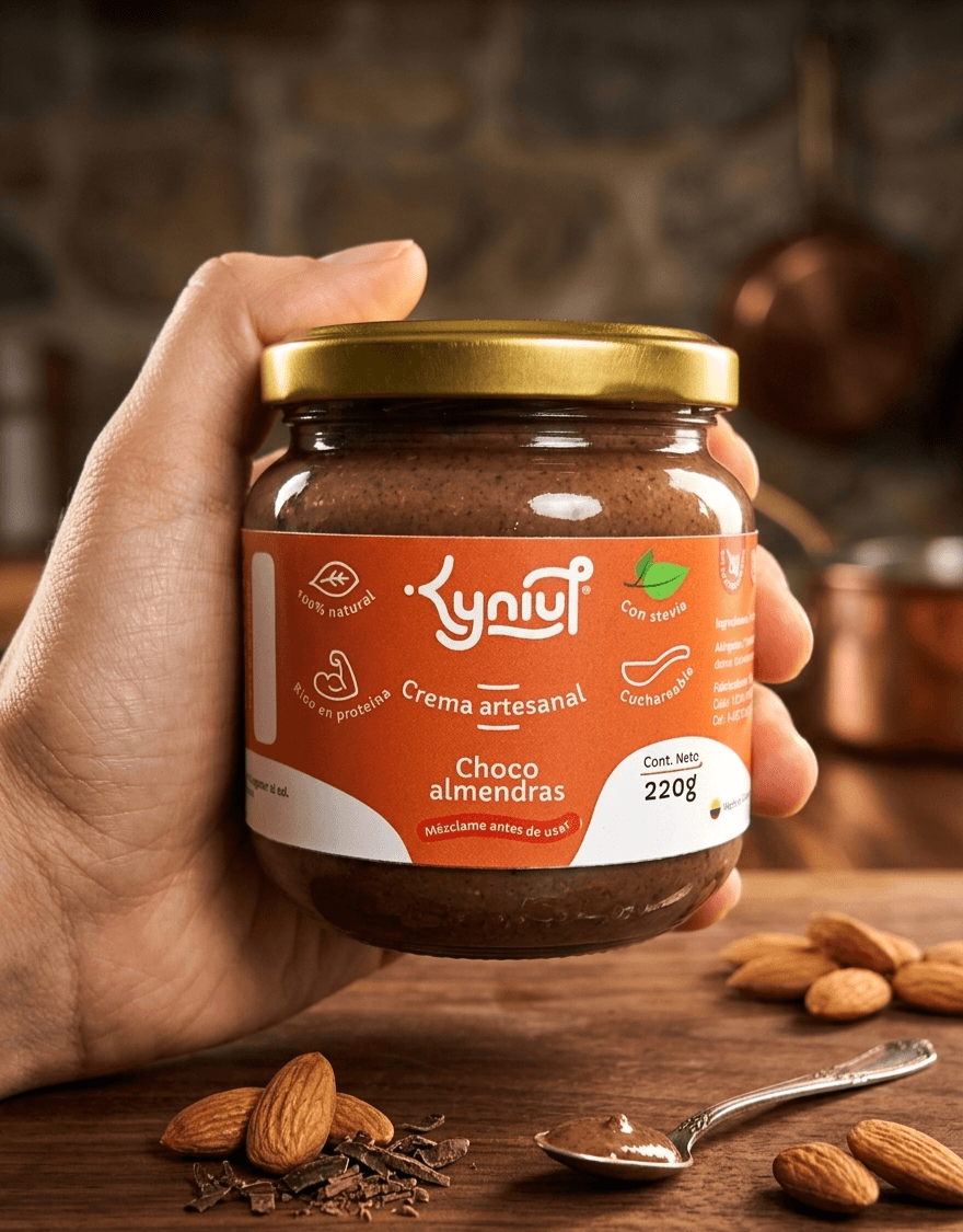 Frasco de Crema artesanal de choco almendras Kyniut de 220g, endulzada con stevia y 100% natural, sostenida por una mano sobre fondo cálido de cocina con almendras enteras y chocolate troceado en una mesa de madera.