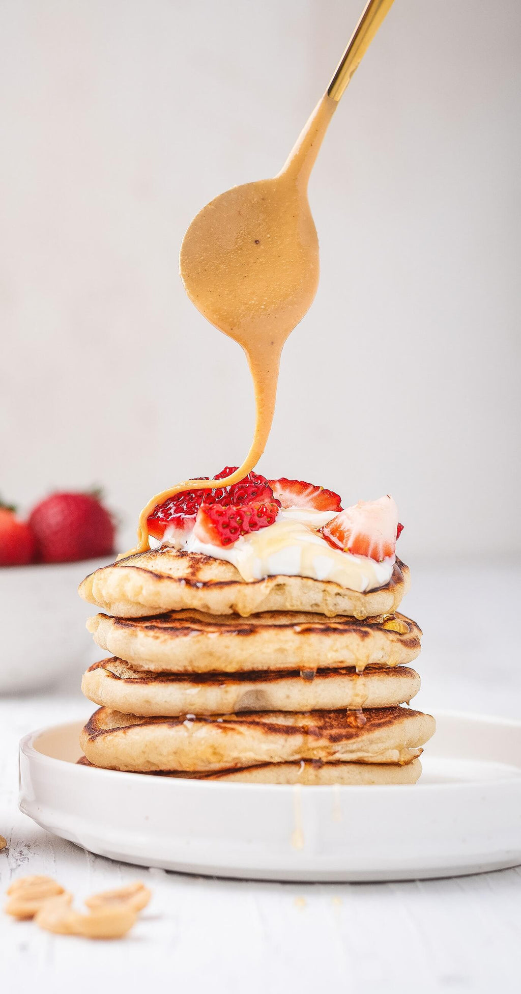 Deliciosa pila de pancakes con fresas y crema, con un chorro de Crema de Maní Kyniut 100% natural cayendo tentadoramente desde una cuchara.