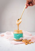 Frasco de Crema de Marañón Kyniut artesanal de 220g, 100% natural y sin azúcar, con una cuchara dorada mostrando su textura suave junto a nueces de marañón.